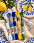Cinco meadas DMC B5200, 799, 820, 973 e 3347 junto a azulejos portugueses e um bastidor com limões bordados em ponto cruz.