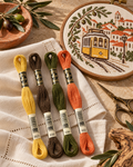 Paleta com meadas DMC 3821, 839, 936 e 922 sobre linho, junto a um elétrico de Lisboa bordado em ponto cruz e ramos de oliveira.