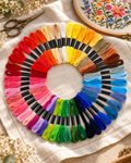 Kit de meadas para bordar em cores variadas, dispostas em círculo sobre linho, junto a um bastidor com flores em ponto cruz.