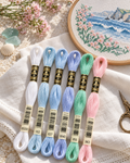 Paleta Primavera Atlântica com seis meadas DMC B5200, 162, 827, 3840, 955 e 963 sobre linho, junto a um bastidor com ondas e flores bordadas em ponto cruz.